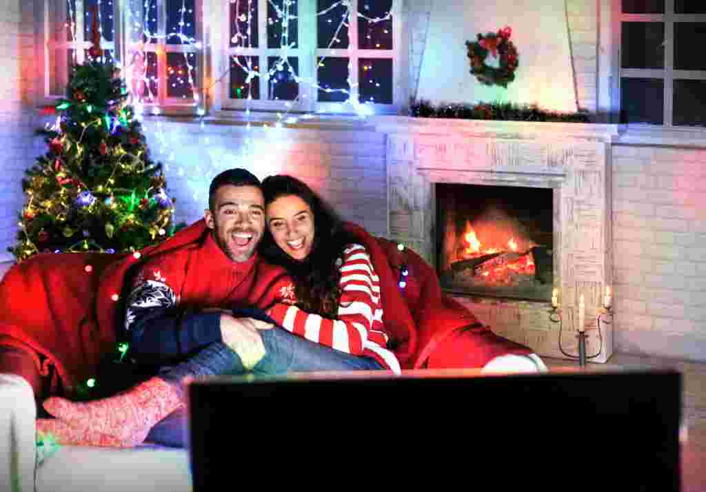 Smiling young couple having fun at home, watching TV together and laughing. Both wears warm clothes and sitting on sofa. On background lights on decorated christmas tree, windows and burning fireplace, steel candlestick on ground. Back of TV set on foreground.